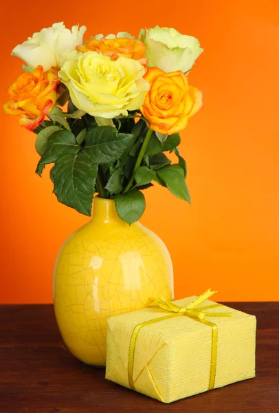 Hermoso ramo de rosas en jarrón con regalo en la mesa sobre fondo naranja — Foto de Stock