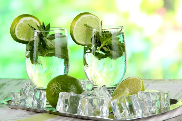 Glasses of cocktail with ice on metal tray on napkin on wooden table on nature background — Stock Photo, Image