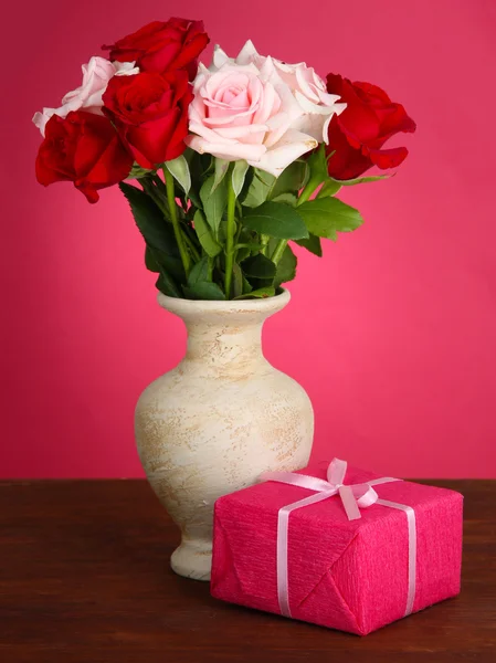 Beautiful bouquet of roses in vase with gift on table on pink background — Stock Photo, Image