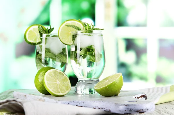 Glasses of cocktail with ice on board on napkin on wooden table on window background — Stock Photo, Image