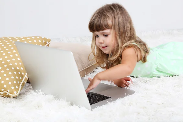Pequena menina bonito encontra-se no tapete com laptop, no fundo cinza — Fotografia de Stock