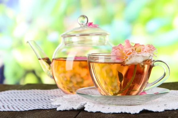 Чайник и чашка чая из чайной розы на салфетке на деревянном столе на природе — стоковое фото