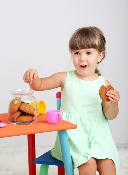 Little cute girl sitting on little chair near table and eating tasty cookie, on gray background — Stock Photo, Image