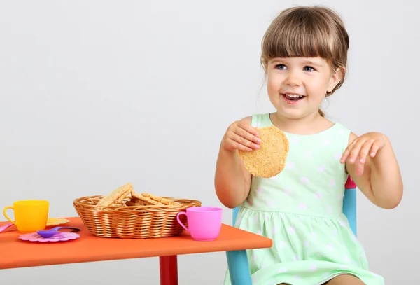 Pequena menina bonito sentado em pequena cadeira perto da mesa e comer biscoito saboroso, no fundo cinza — Fotografia de Stock