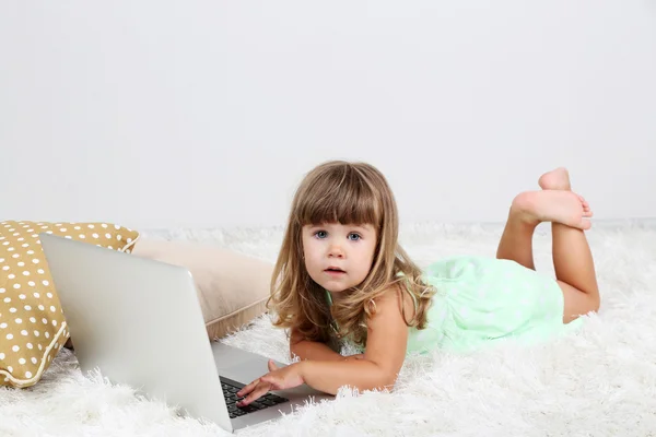 Pequena menina bonito encontra-se no tapete com laptop, no fundo cinza — Fotografia de Stock