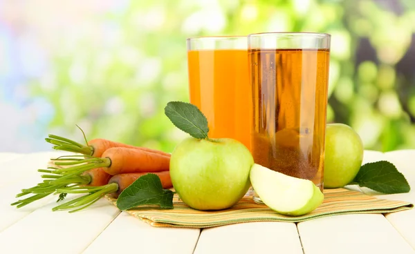 Glazen van SAP, appelen en wortelen op witte houten tafel, op groene achtergrond — Stockfoto