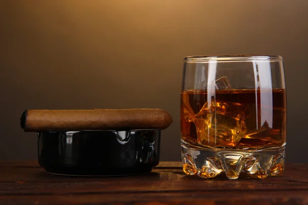 Brandy glass with ice and cigar on wooden table on brown background — Stock Photo, Image