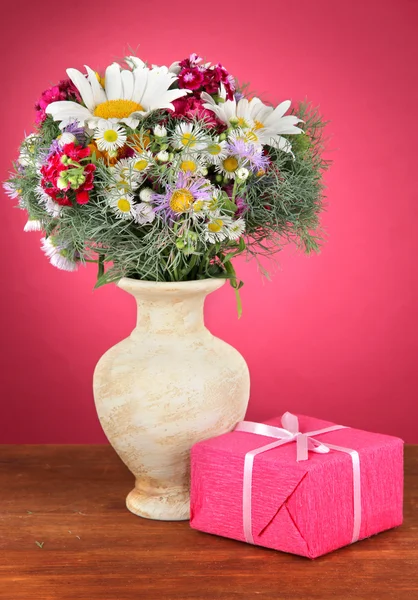 Beautiful bright flowers in vase with gift on table on pink background — Stock Photo, Image