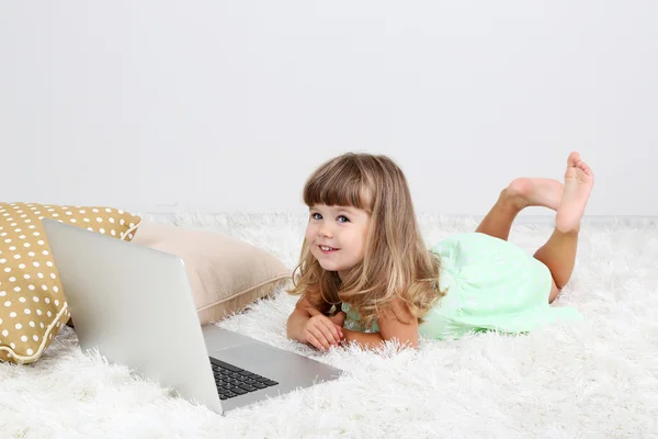 Pequena menina bonito encontra-se no tapete com laptop, no fundo cinza — Fotografia de Stock