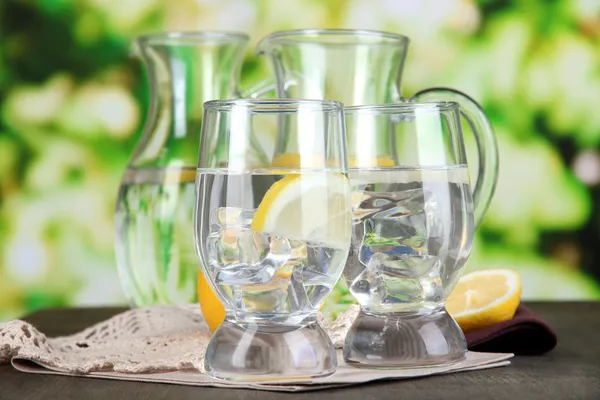 Jarras de vidrio de agua y vasos sobre mesa de madera sobre fondo natural —  Fotos de Stock