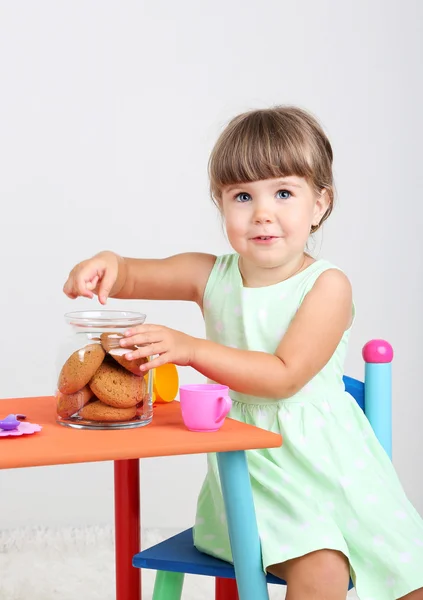 Pequena menina bonito sentado em pequena cadeira perto da mesa e comer biscoito saboroso, no fundo cinza — Fotografia de Stock