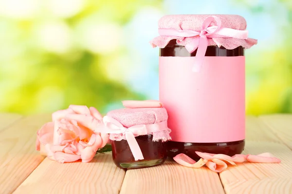 Vasi con marmellata di rose e fiori su tavolo di legno su sfondo verde — Foto Stock