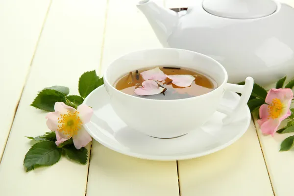 Xícara e bule de chá de ervas com flores de rosa do quadril na mesa de madeira branca — Fotografia de Stock