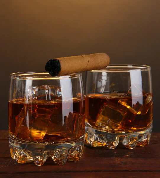 Brandy glasses with ice and cigar on wooden table on brown background — Stock Photo, Image