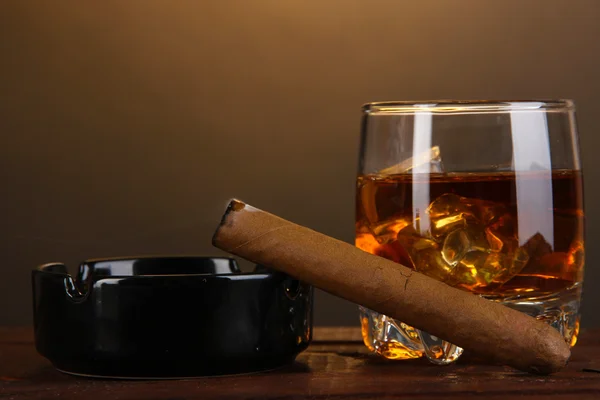 Brandy glass with ice and cigar on wooden table on brown background — Stock Photo, Image