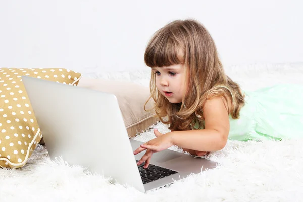 Pequena menina bonito encontra-se no tapete com laptop, no fundo cinza — Fotografia de Stock