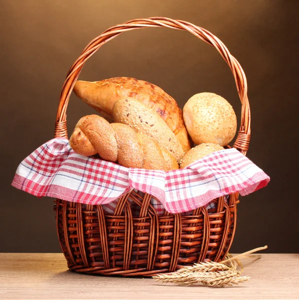 Delicious breads in basket and ears on wooden table on brown background — Stock Photo, Image