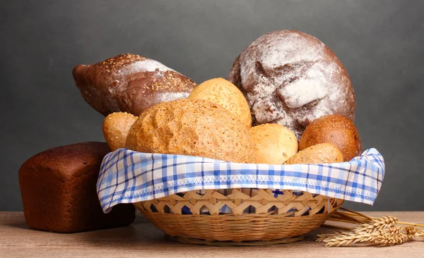 Delicious bread in basket and ears on wooden table on gray background — Stock Photo, Image