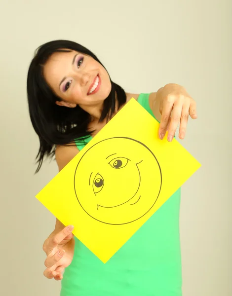 Young woman holding paper with happy smiley on gray background. — Stock Photo, Image