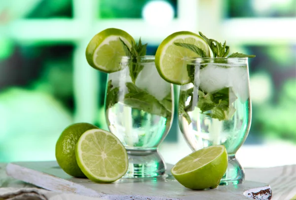 Glasses of cocktail with ice on board on napkin on wooden table on window background — Stock Photo, Image