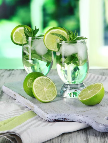 Glasses of cocktail with ice on board on napkin on wooden table on window background — Stock Photo, Image