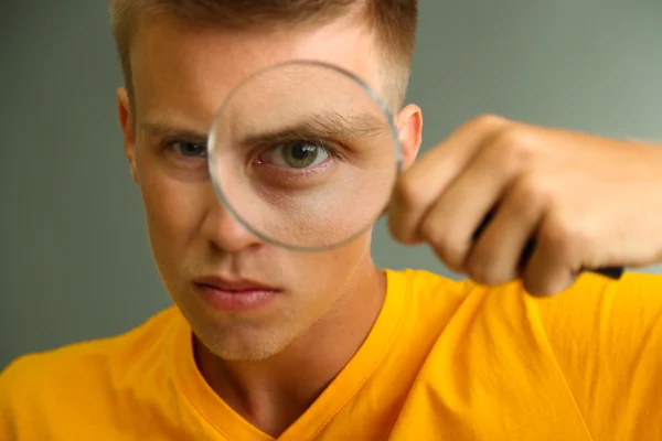 Ung man tittar genom förstoringsglas på grå bakgrund — Stockfoto