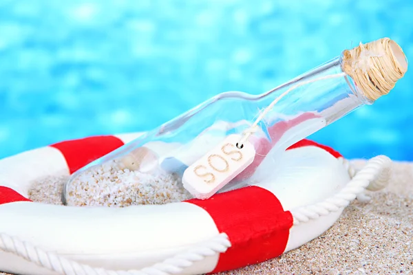 Glass of bottle with note inside on sand, on bright blue background — Stock Photo, Image