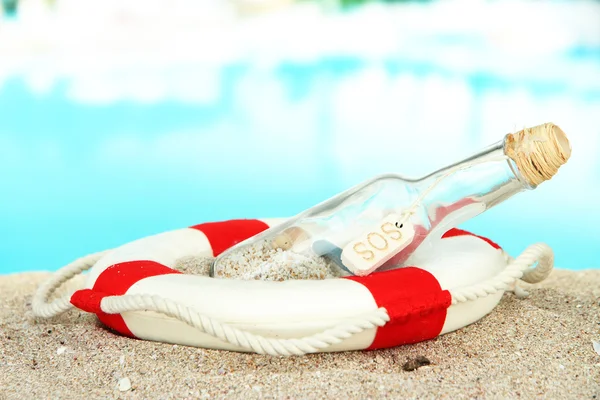 Glazen fles met opmerking binnen op zand, op heldere blauwe achtergrond — Stockfoto