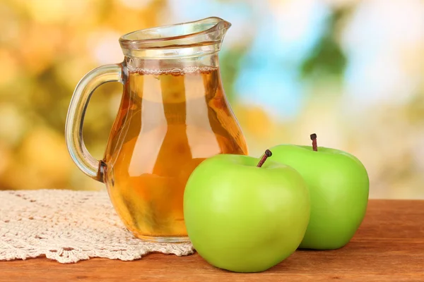 Full jug of apple juice and apple on wooden table on bright background — Stock Photo, Image