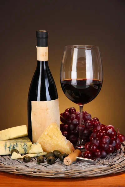 Refined still life of wine, cheese and grapes on wicker tray on wooden table on brown background — Stock Photo, Image