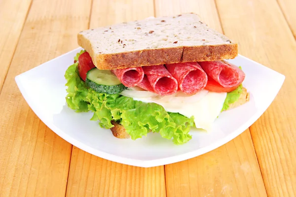 Tasty sandwich with salami sausage and vegetables on white plate, on wooden background — Stock Photo, Image