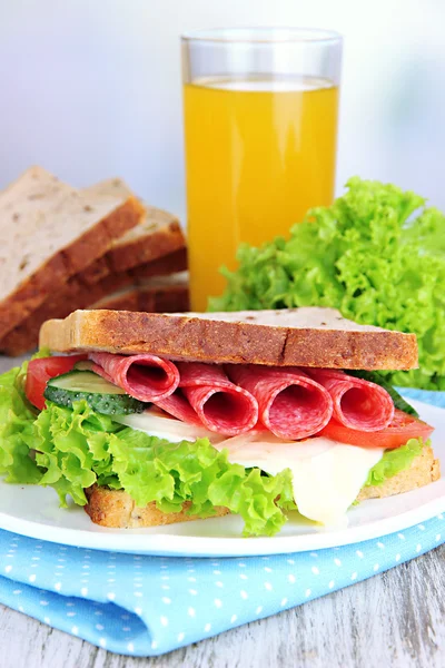 Composition with fruit juice and tasty sandwich with salami sausage and vegetables on color napkin, on wooden table, on bright background — Stock Photo, Image