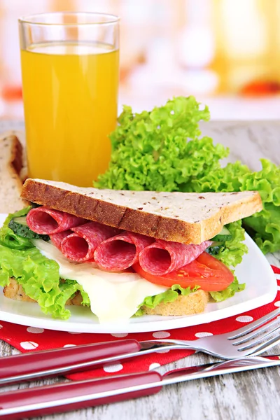 Composition with fruit juice and tasty sandwich with salami sausage and vegetables on color napkin, on wooden table background — Stock Photo, Image