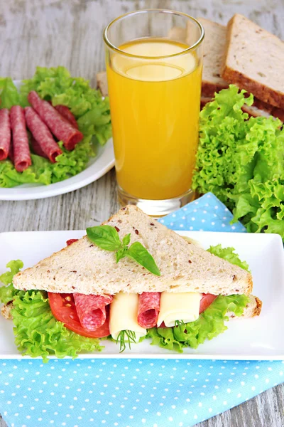 Composición con zumo de fruta y sabroso sándwich con salchicha salami y verduras en servilleta de color, sobre fondo de mesa de madera —  Fotos de Stock