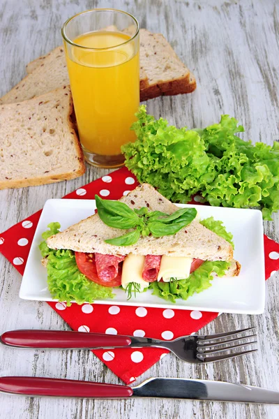 Composition with fruit juice and tasty sandwich with salami sausage and vegetables on color napkin, on wooden table background — Stock Photo, Image