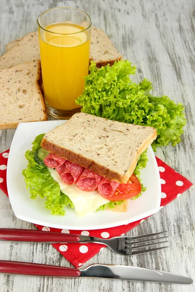 Composition with fruit juice and tasty sandwich with salami sausage and vegetables on color napkin, on wooden table background — Stock Photo, Image