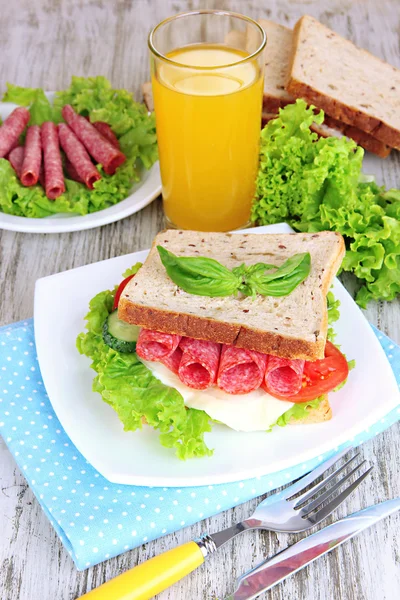 Композиция с фруктовым соком и вкусный сэндвич с колбасой салями и овощами на цветной салфетке, на деревянном фоне стола — стоковое фото
