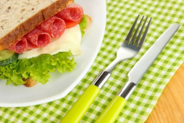 Tasty sandwich with salami sausage and vegetables on white plate, on wooden background — Stock Photo, Image