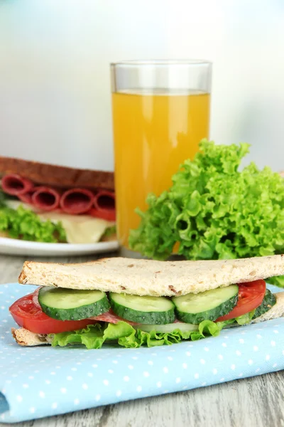 Composition with fruit juice and tasty sandwich with salami sausage and vegetables on color napkin, on wooden table, on bright background — Stock Photo, Image