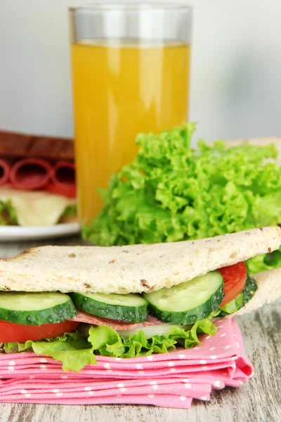 Composition with fruit juice and tasty sandwich with salami sausage and vegetables on color napkin, on wooden table, on bright background — Stock Photo, Image