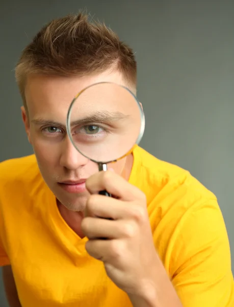 Young man looking through magnifying glass on grey background — Stock Photo, Image