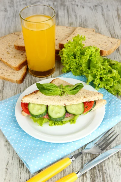 Composition with fruit juice and tasty sandwich with salami sausage and vegetables on color napkin, on wooden table background — Stock Photo, Image