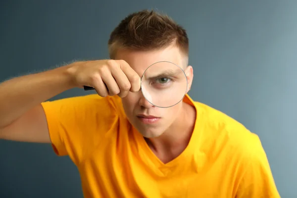 Young man looking through magnifying glass on grey background — Stock Photo, Image