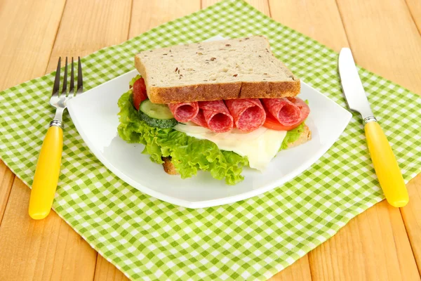 Tasty sandwich with salami sausage and vegetables on white plate, on wooden background — Stock Photo, Image