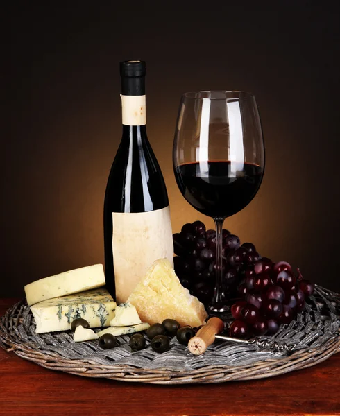 Refined still life of wine, cheese and grapes on wicker tray on wooden table on brown background — Stock Photo, Image