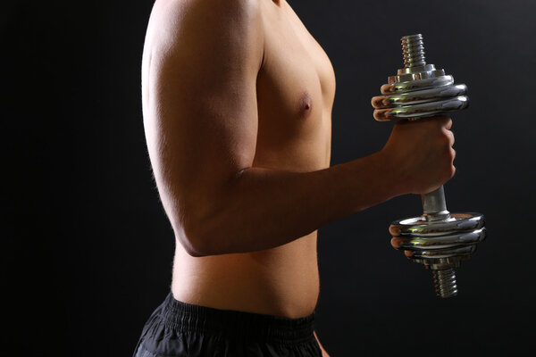Handsome young muscular sportsman execute exercise with dumbbell on dark background