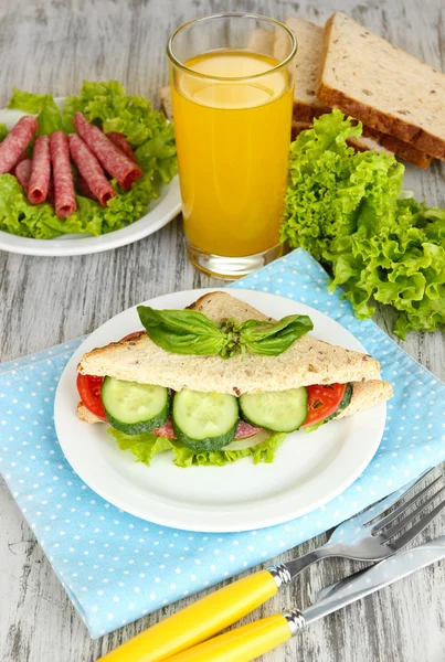 作文与果汁和美味三明治与萨拉米香肠和蔬菜上彩色餐巾，木制表背景上 — 图库照片