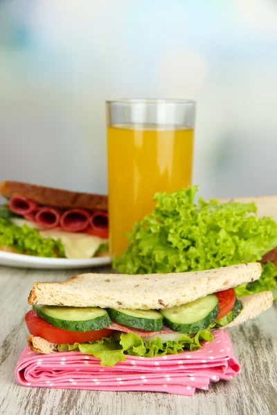 Composition with fruit juice and tasty sandwich with salami sausage and vegetables on color napkin, on wooden table, on bright background — Stock Photo, Image