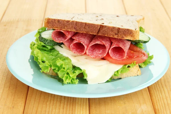 Tasty sandwich with salami sausage and vegetables on blue plate, on wooden background — Stock Photo, Image