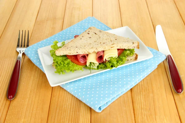 Вкусный сэндвич с колбасой и овощами на белой тарелке, на деревянном фоне — стоковое фото